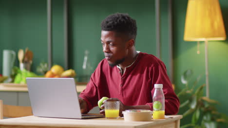Joven-Negro-Comiendo-Manzana-Y-Usando-Una-Laptop-En-Casa